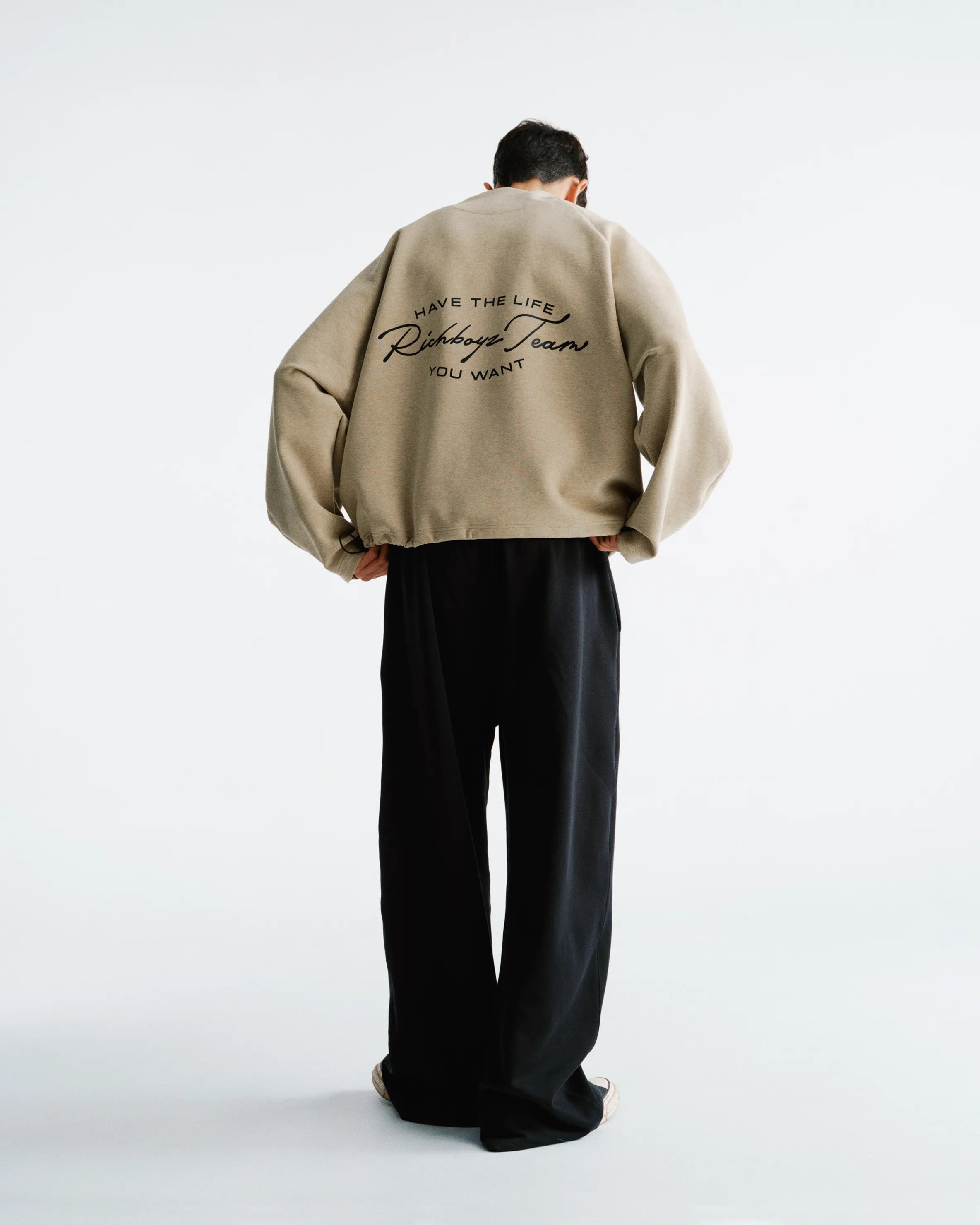 Person wearing a beige jacket with text on the back, standing against a white background